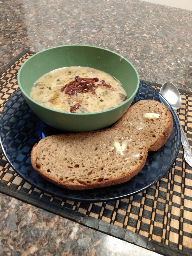 A finished bowl of clam chowder, topped with bits of bacon and set on a plate next to two slices of homemade brown bread.