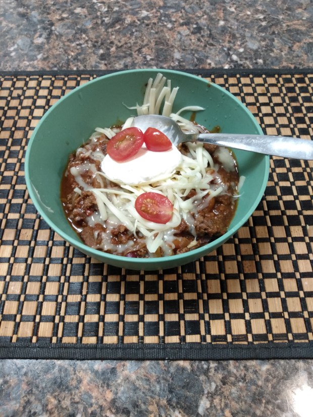 The chili, in a bowl, topped with shredded cheddar, sour cream and tomatoes.