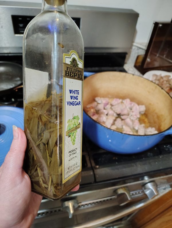 In the foreground, half a bottle of white wine vinegar stuffed with drab-looking tarragon. In the background, my blue Dutch oven with cubes of pork browning on the stove.